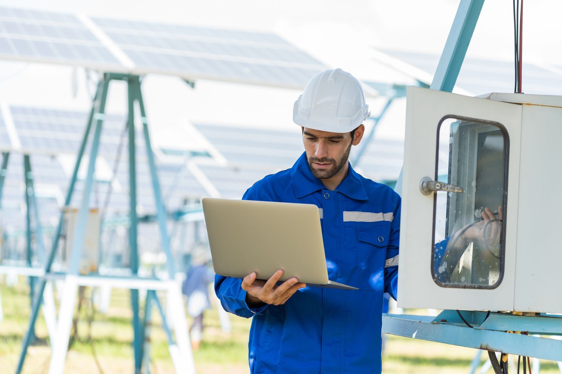 Technician in blue coveralls and hard hat uses laptop near solar panels and control box.