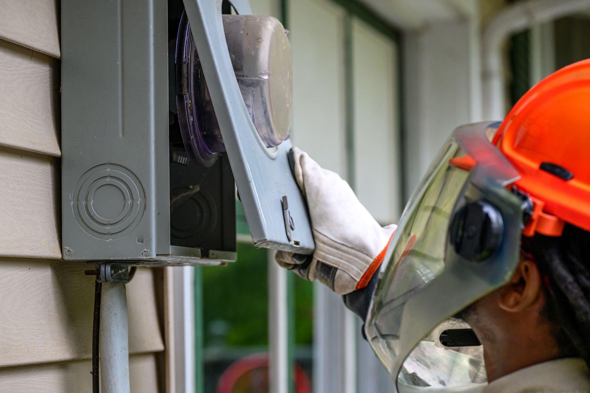 Electrician in safety gear working on a gray electrical box on a beige house exterior.