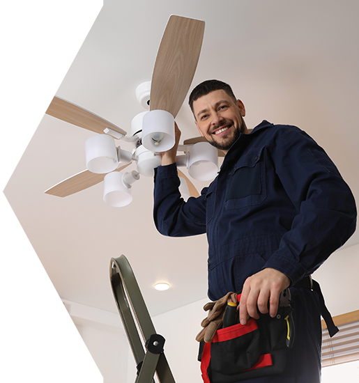 Electrician installing a ceiling fan, standing on a ladder. He is smiling and wearing a blue uniform.