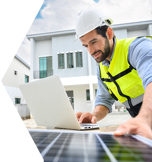 Man in hard hat and safety vest working on laptop, solar panel in foreground, house in background.