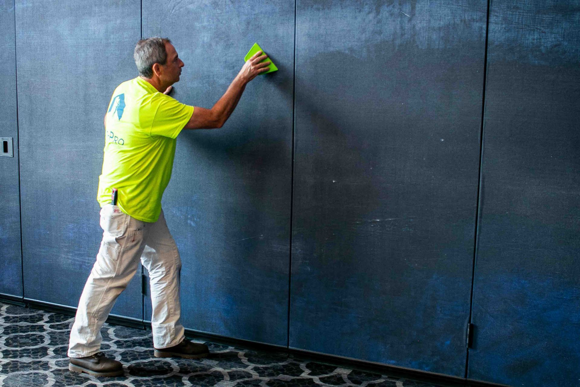 Man in yellow shirt using a squeegee on a dark-colored wall indoors.