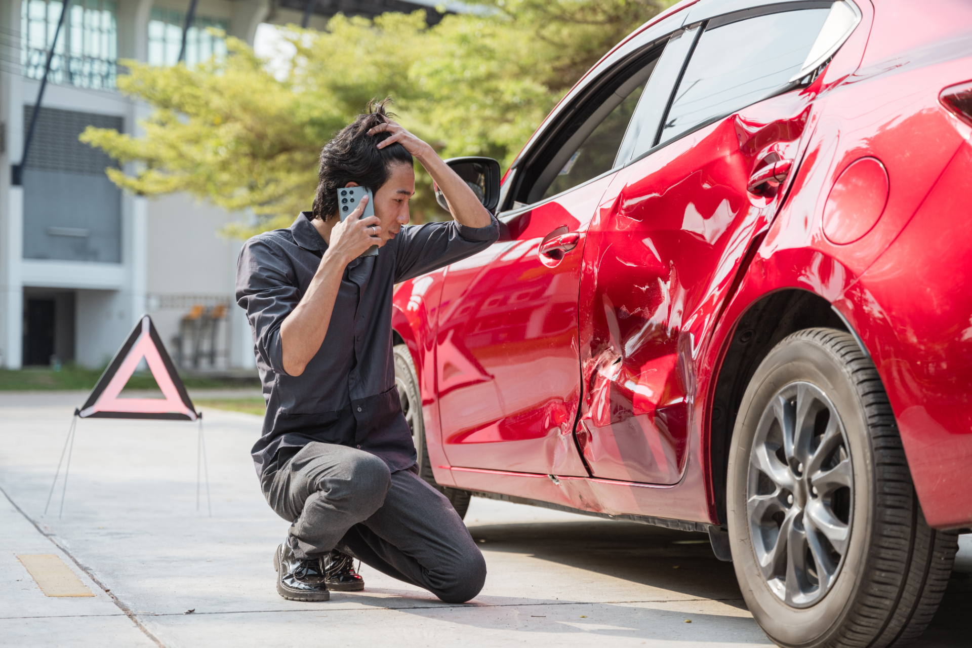 Man kneeling by damaged red car, holding phone, looking distressed. Emergency triangle on ground.