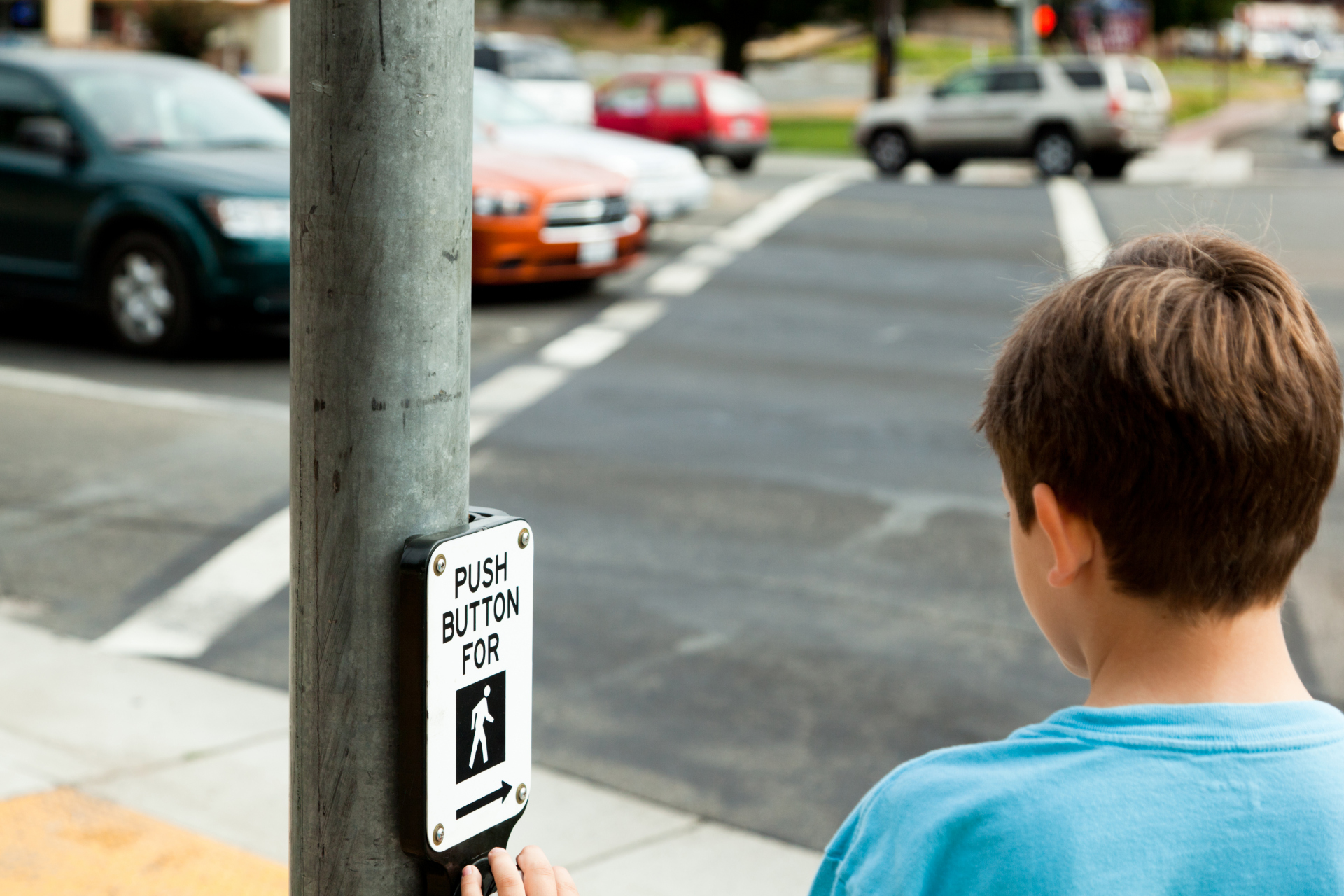 Boy at crosswalk button, waiting to cross street with cars in background.