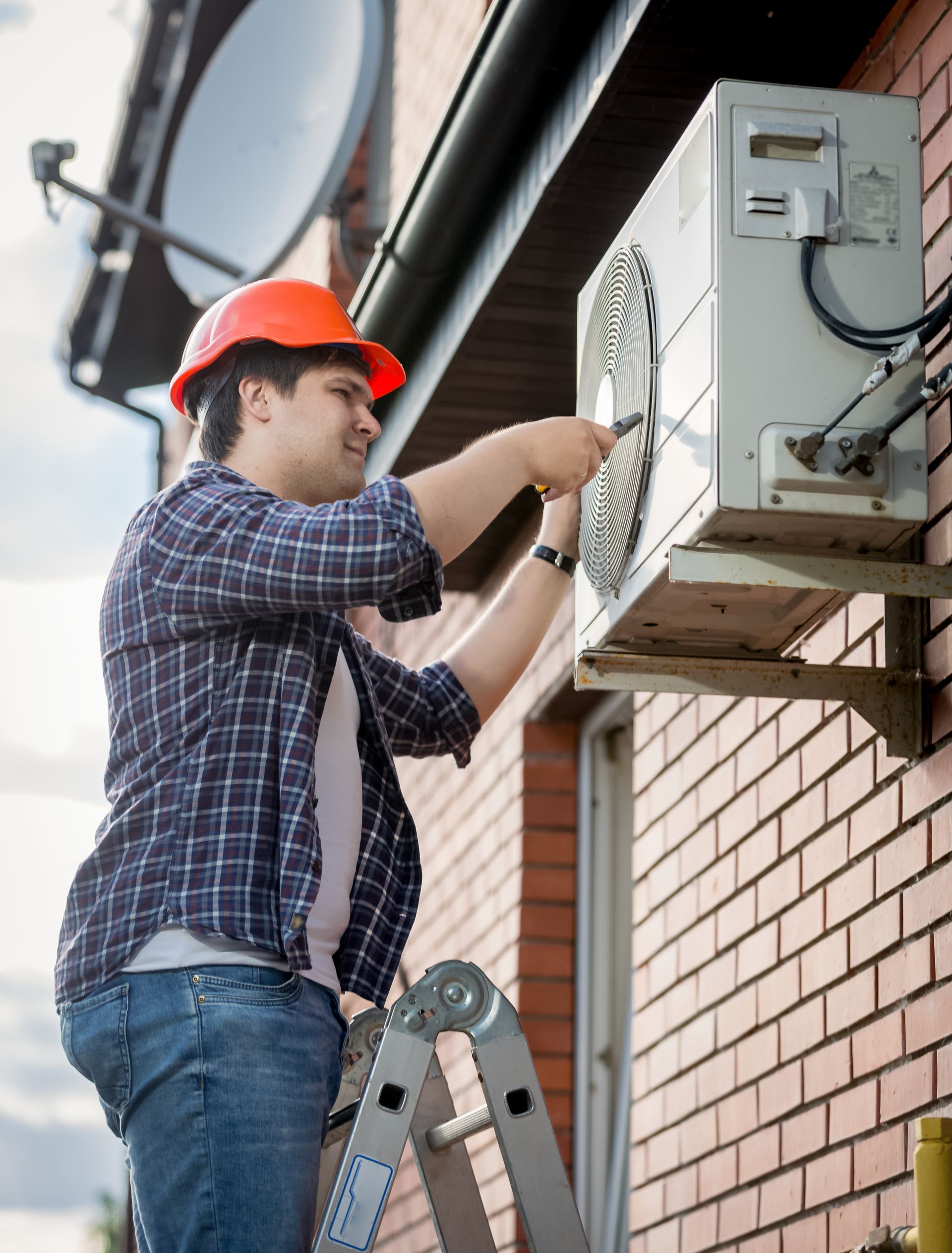 A man is standing on a ladder fixing an air conditioner on the side of a building.