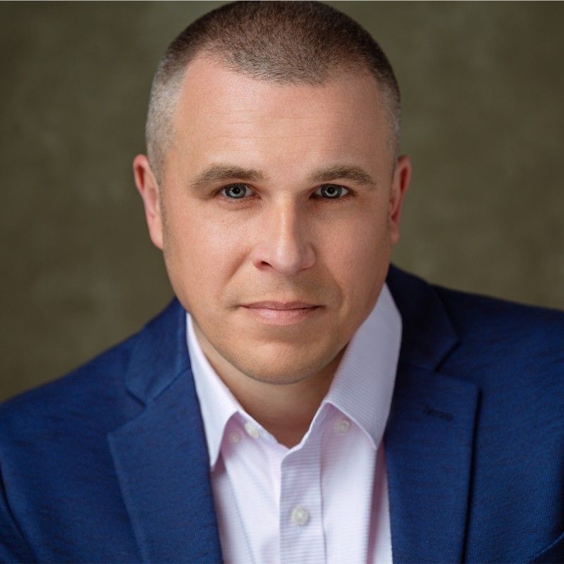 Man in a blue blazer and pink shirt, looking at the camera. Neutral background.