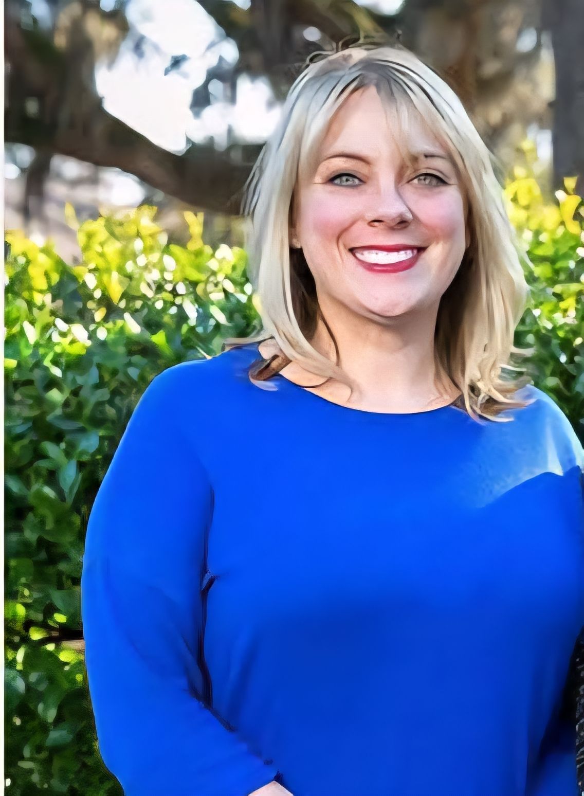 Woman with blonde hair, smiling in a blue top outdoors with green foliage in the background.