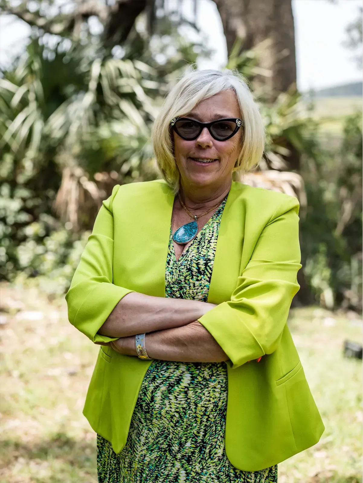 Woman wearing sunglasses and lime green blazer, arms crossed, standing outdoors.