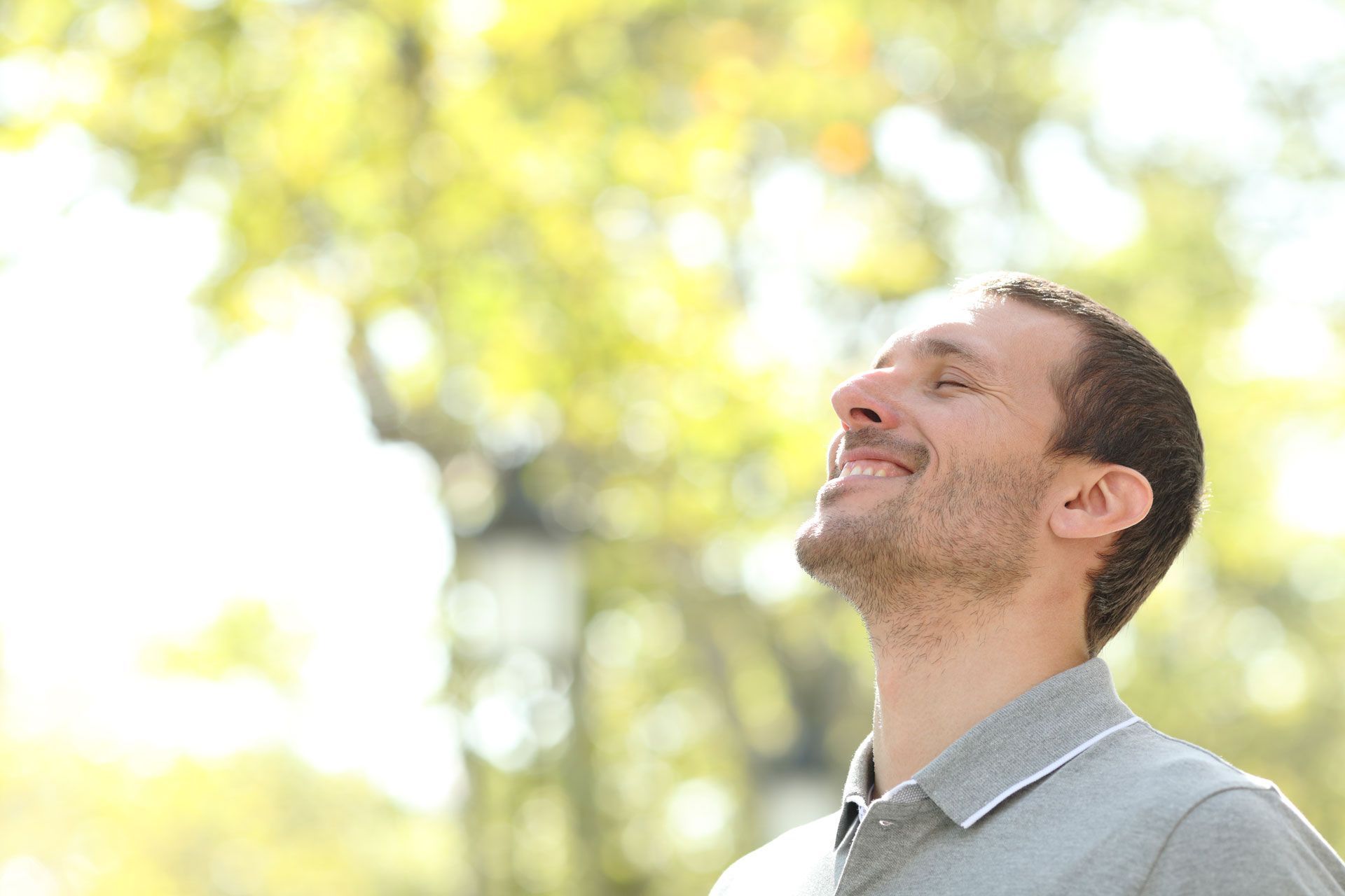Man looking up, smiling, outside with trees in the background, enjoying the sunshine.