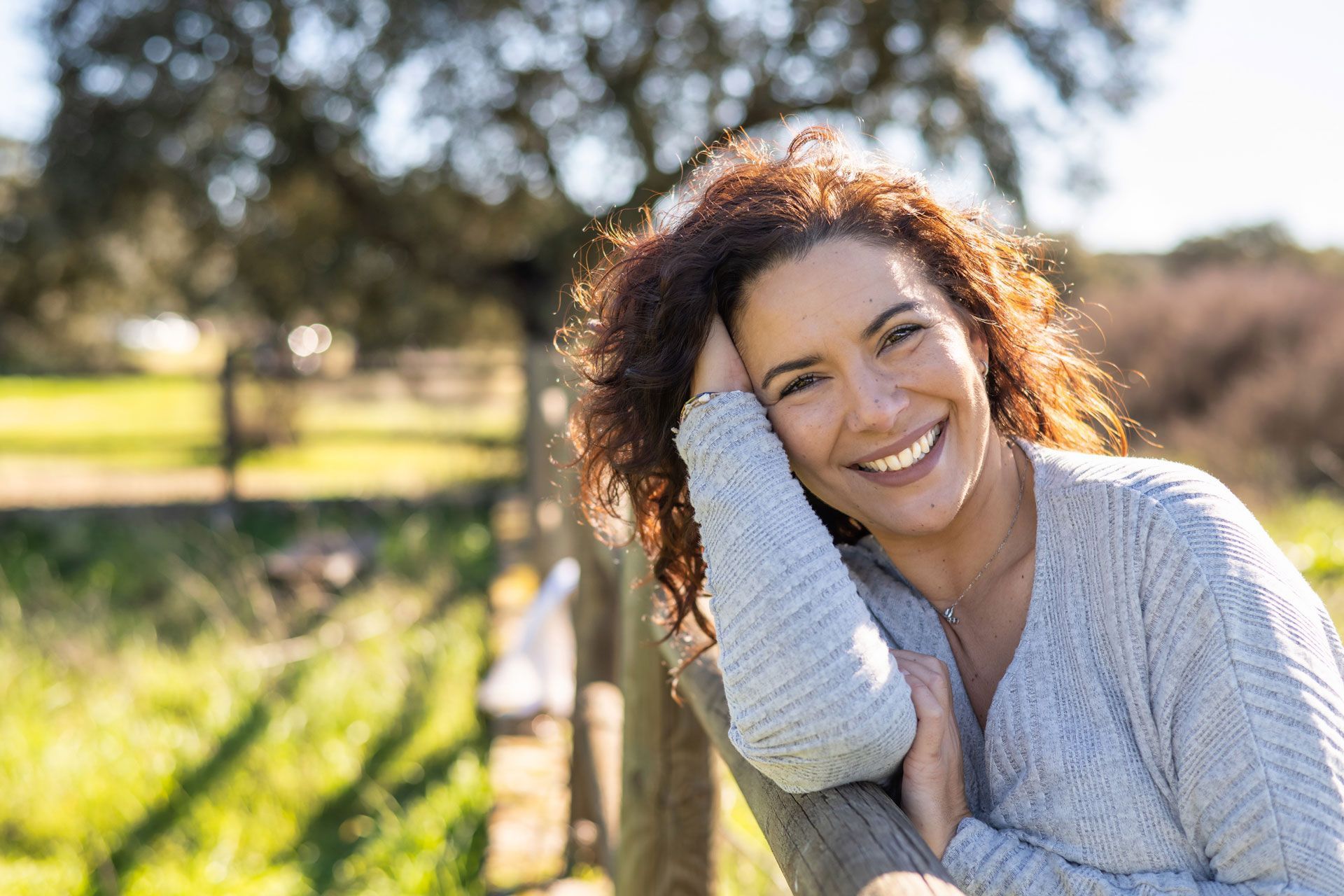 Woman leaning on a wooden fence, smiling outdoors. Curly brown hair, light blue sweater, sunny day.