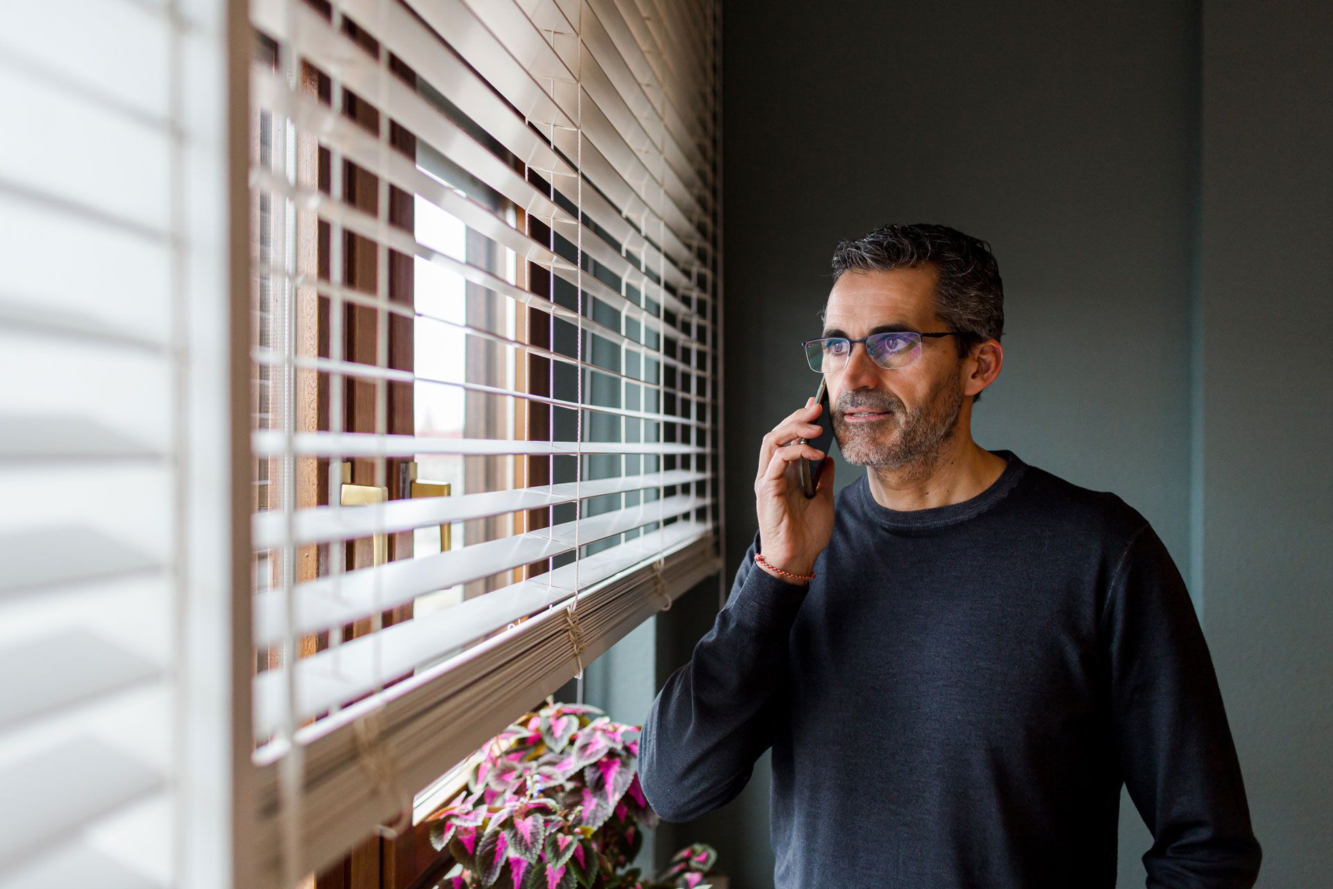 Man with glasses on a phone call near a window with blinds.