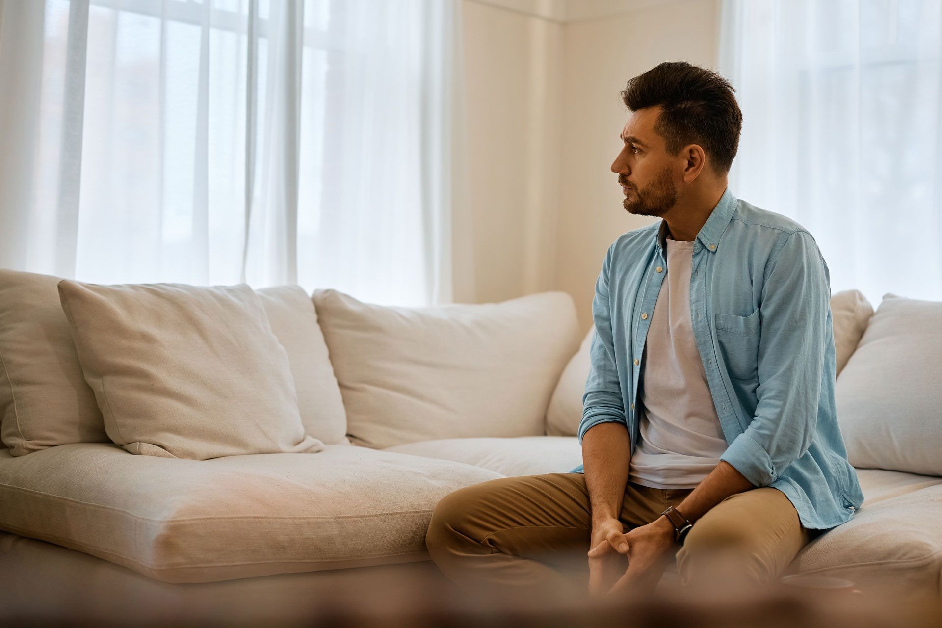 Man seated on a white sofa, looking to the side. Wearing a blue shirt and khaki pants, indoors.