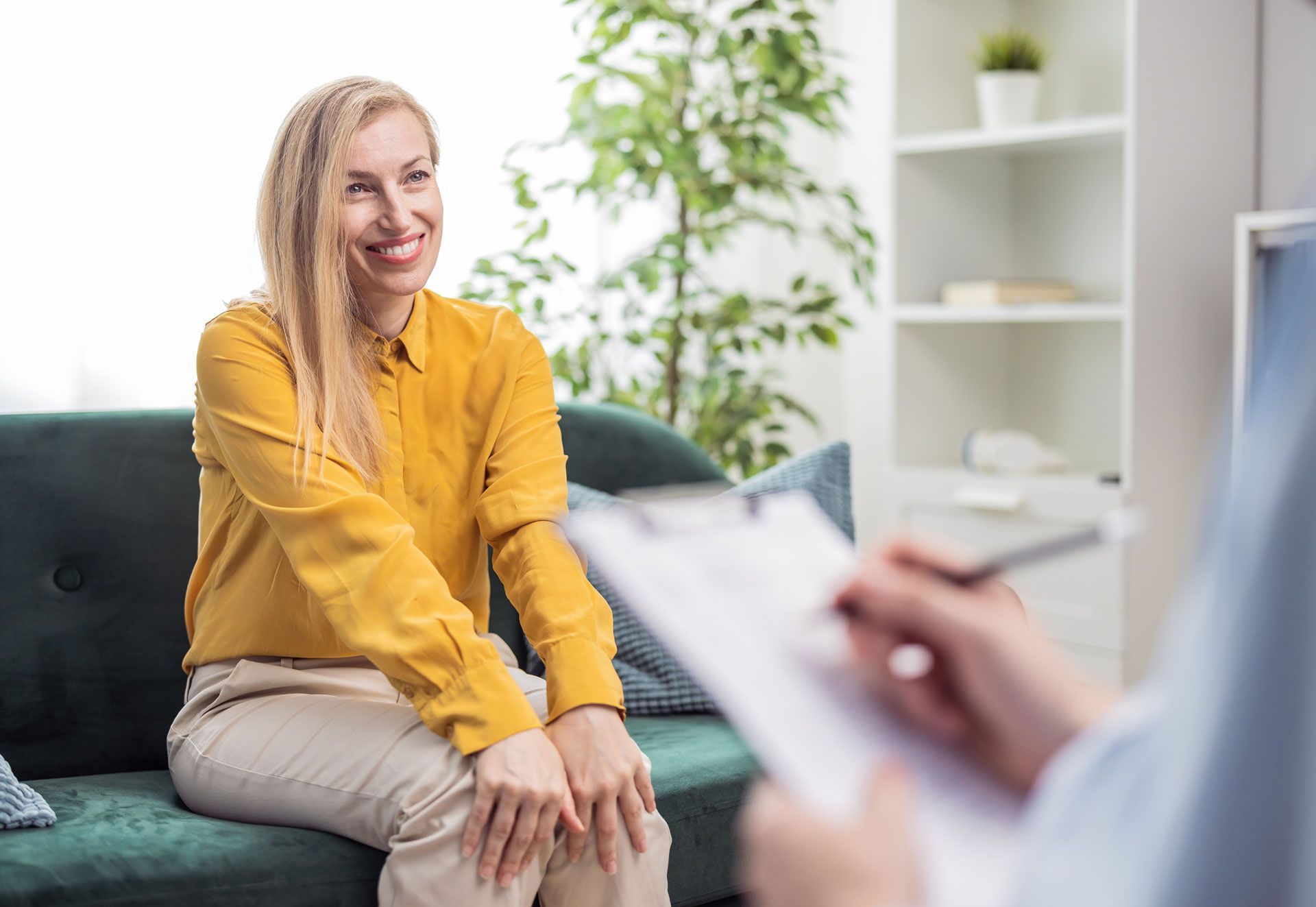 Woman in yellow shirt smiles, sitting on a green couch, facing person writing on clipboard. Bright room setting.
