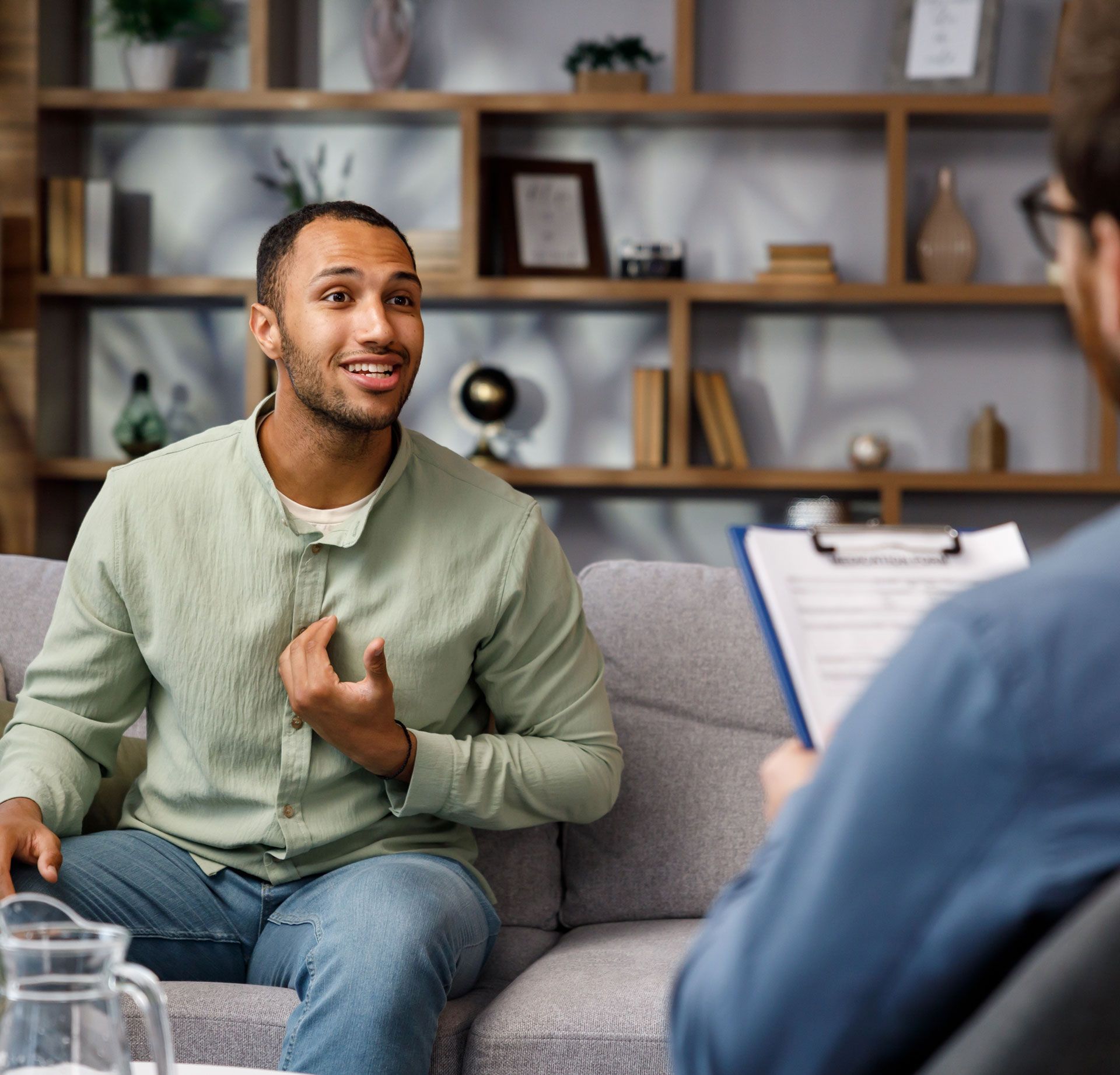 Man on couch talking to person holding clipboard, in a room with a bookshelf.