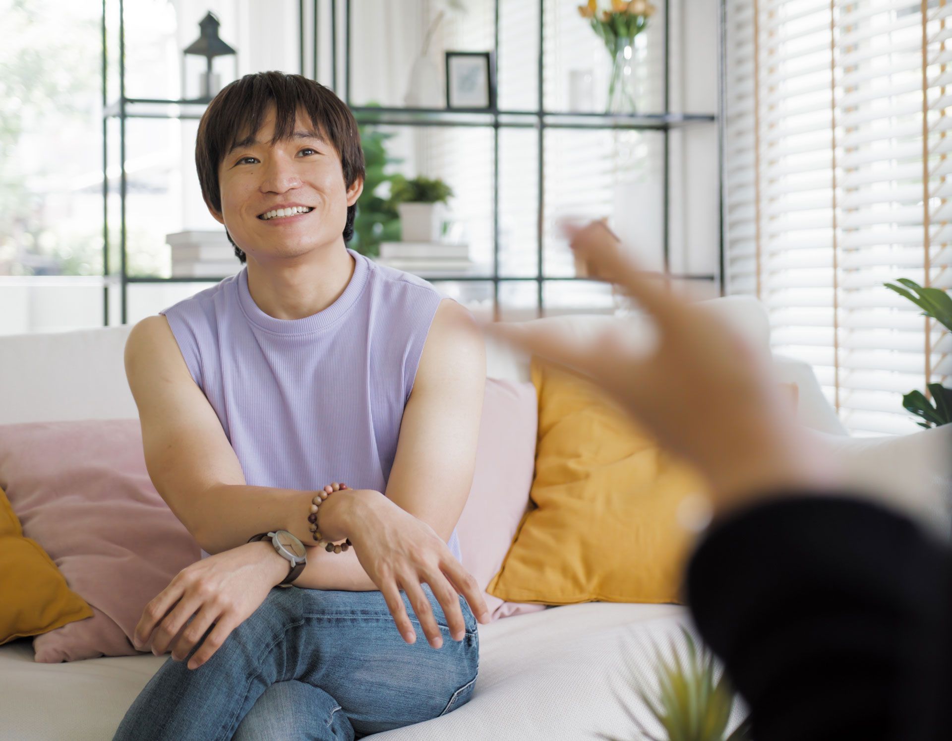 Man in lilac top and jeans smiles while gesturing, seated on a couch indoors.