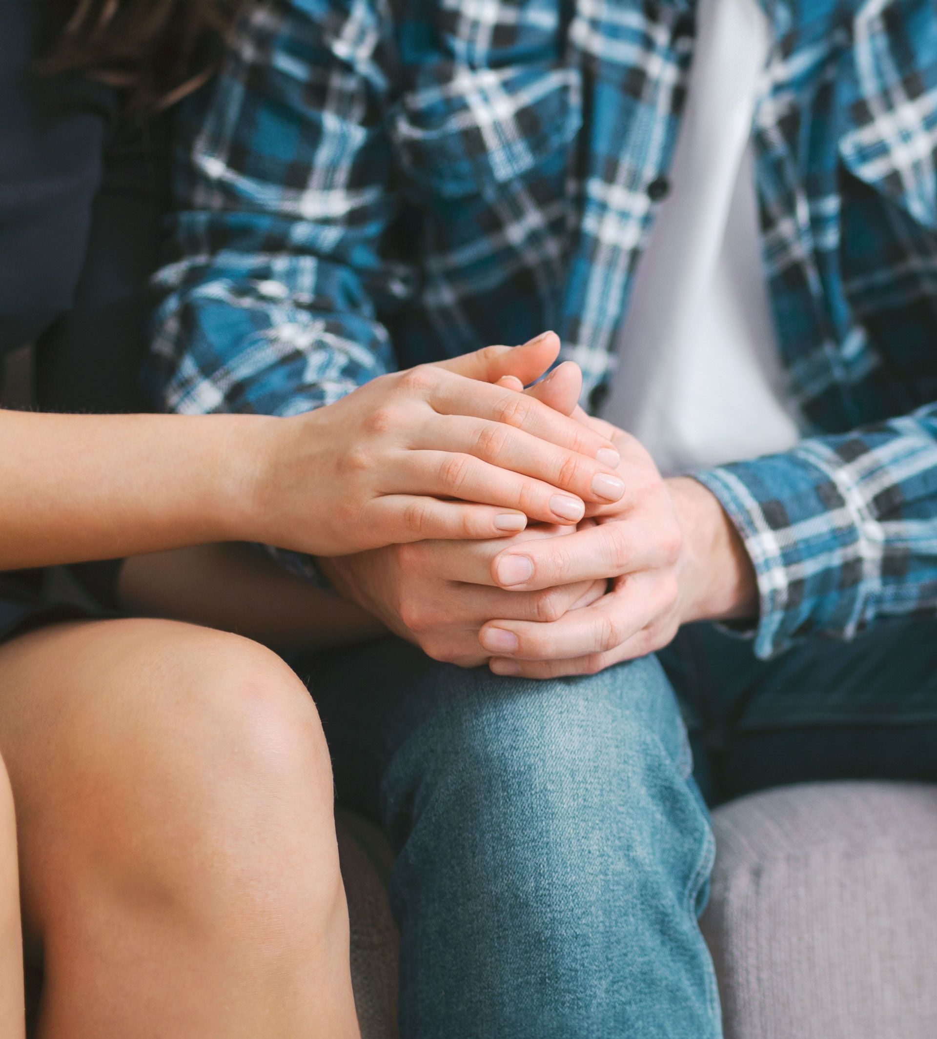 Person's hands clasped together, seated. One person in blue plaid shirt, other in white shirt.