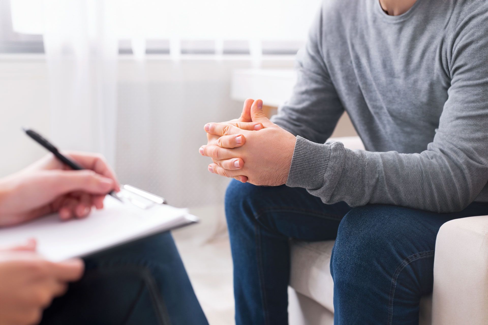 Therapist taking notes during a session with a person sitting on a couch, hands clasped, light room.