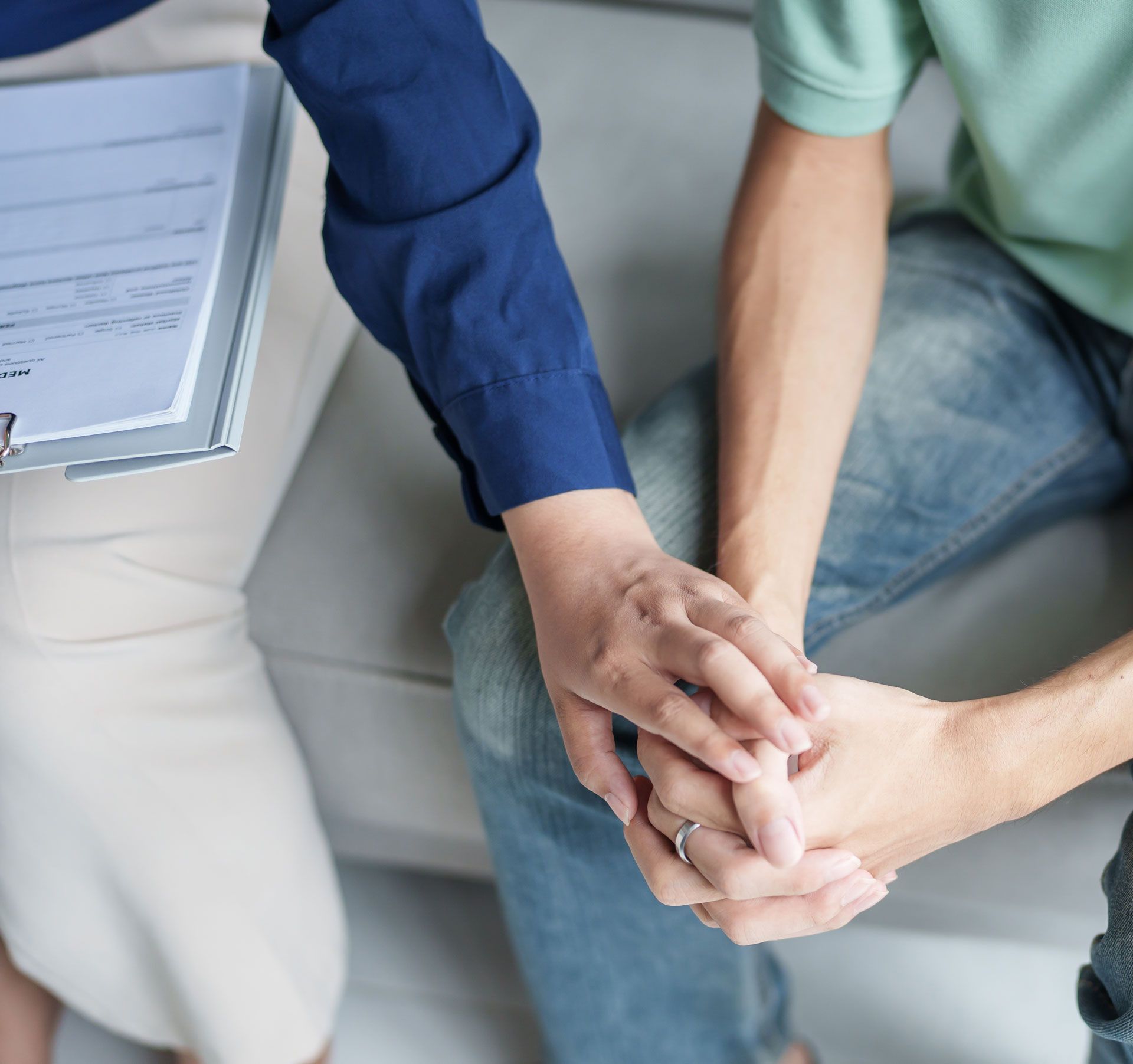 Person's hands held by another's, on a couch. One person wears jeans and a green shirt.