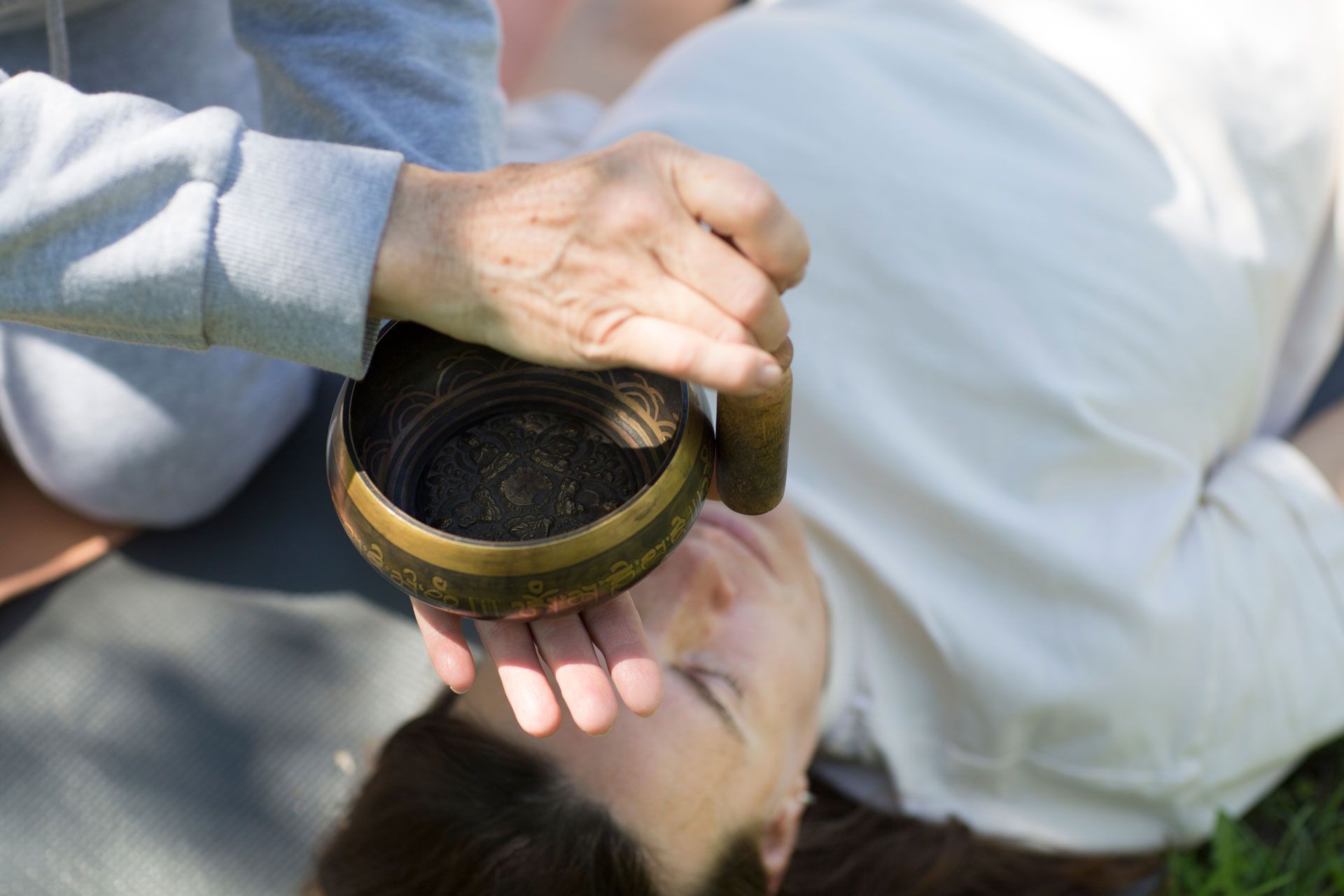Person receiving sound therapy. A hand holds a singing bowl near a person's face outdoors.