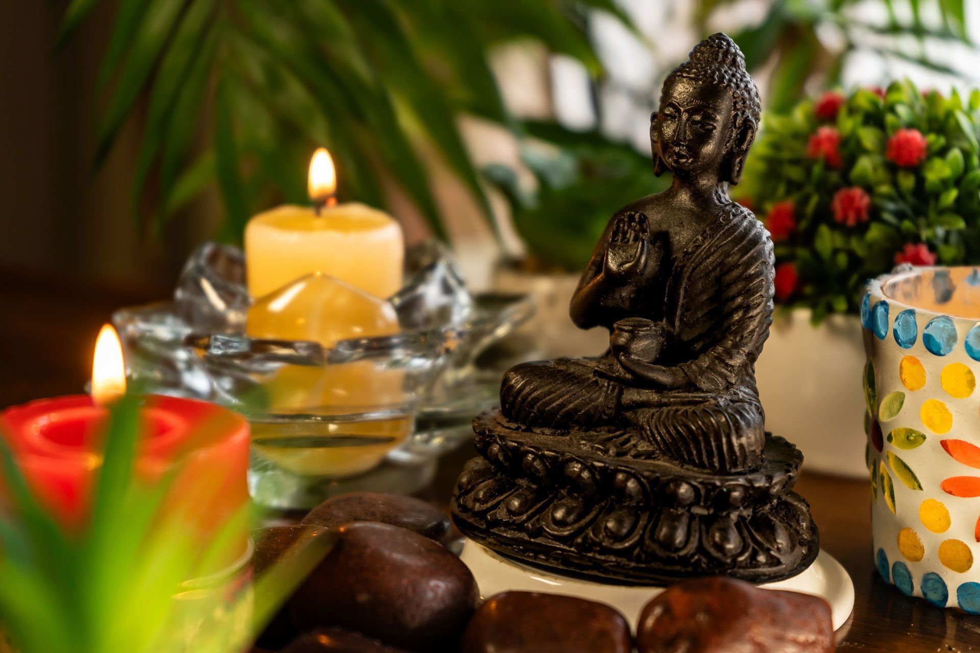 Buddha statue in meditation pose, lit by candles and surrounded by plants and stones.