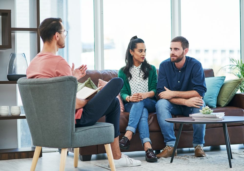 Therapist talking to a couple on a couch in a well-lit living room.