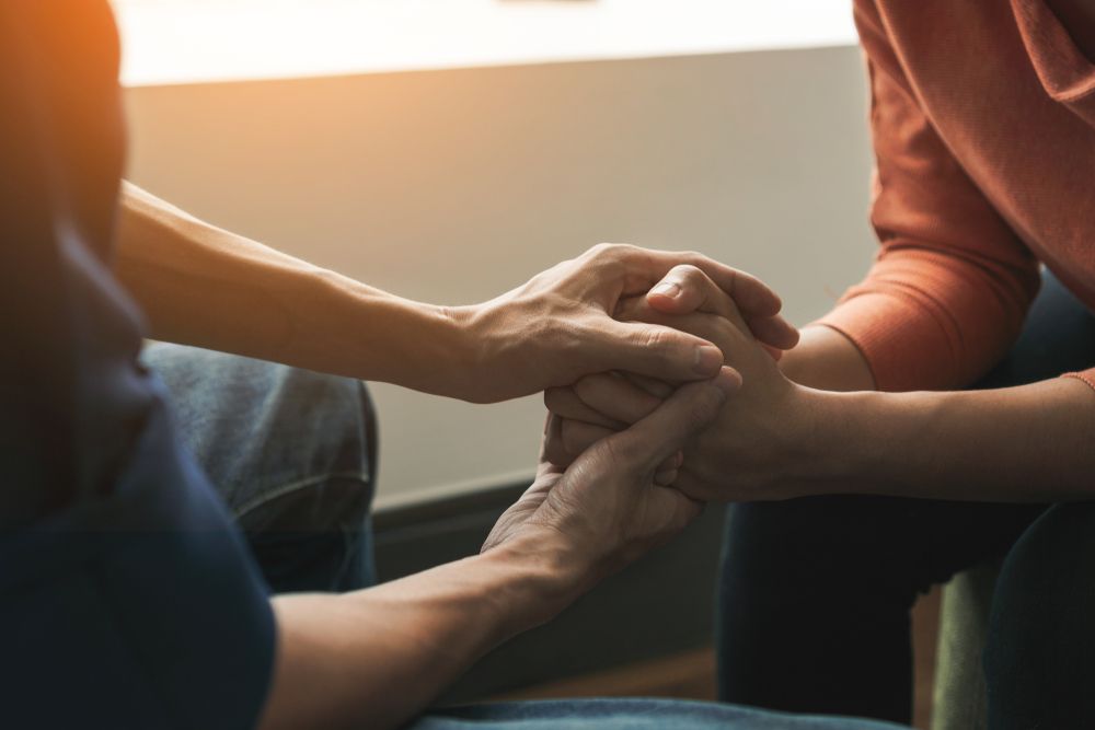 Hands clasped in support, warm lighting, seated indoors.