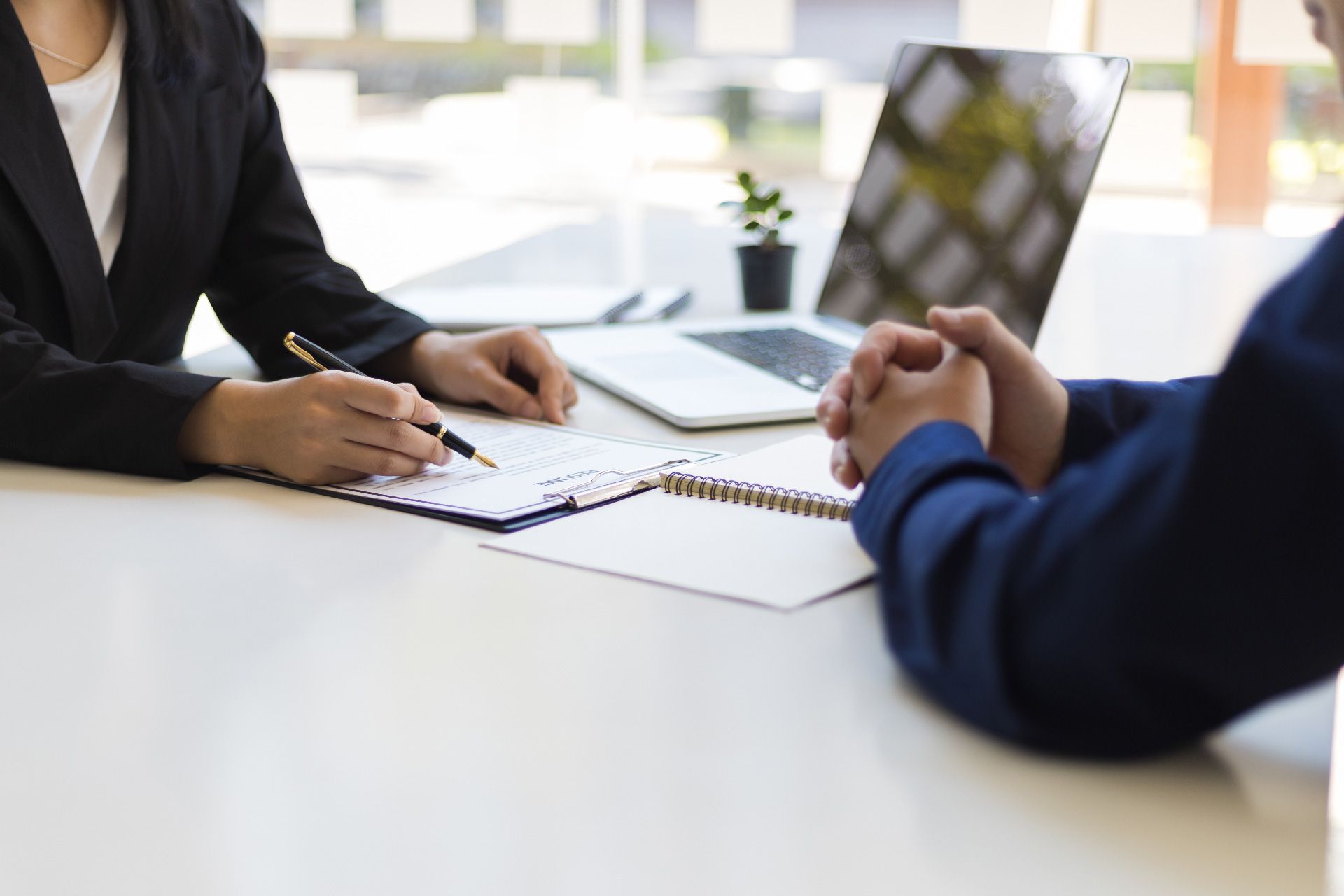 Dos personas vestidas de negocios sentadas en una mesa, una escribiendo en un portapapeles y la otra con las manos entrelazadas, probablemente en una entrevista.
