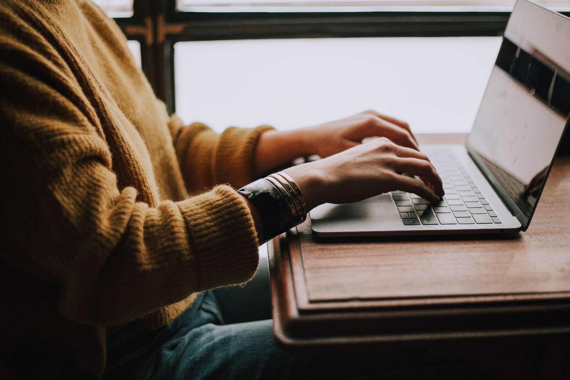 Persona escribiendo en una computadora portátil en un escritorio de madera, vistiendo un suéter amarillo y sentada junto a una ventana.