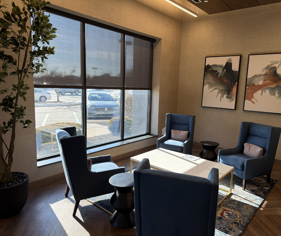 A sunlit office waiting area featuring four blue wingback chairs, a central coffee table, and two abstract wall paintings.