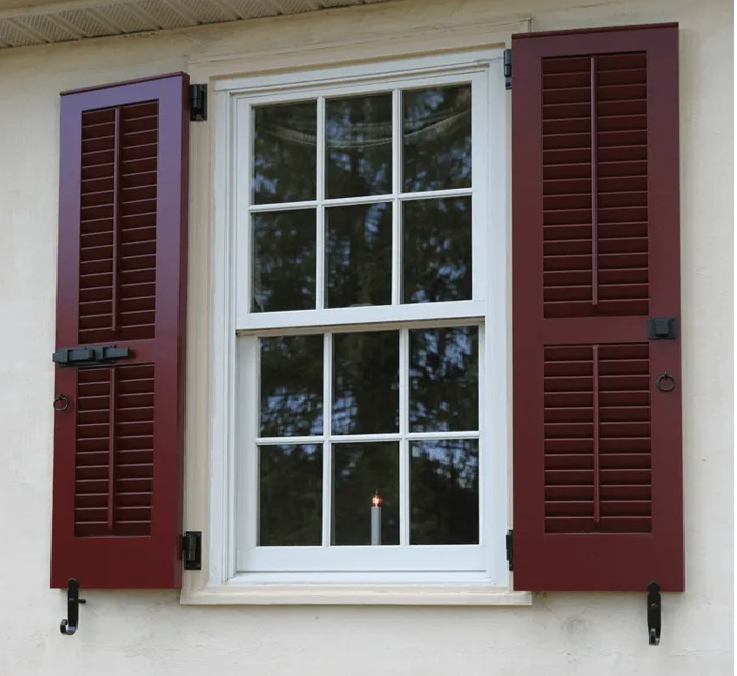 A white window with six-pane glass divided into two sashes, flanked by dark red louvered shutters and black iron hardware.