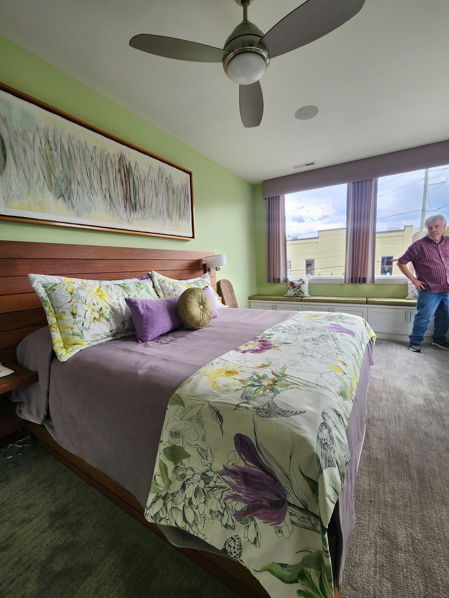 A bedroom features a large wood headboard, a bed with floral bedding, light green walls, a ceiling fan, and a person.