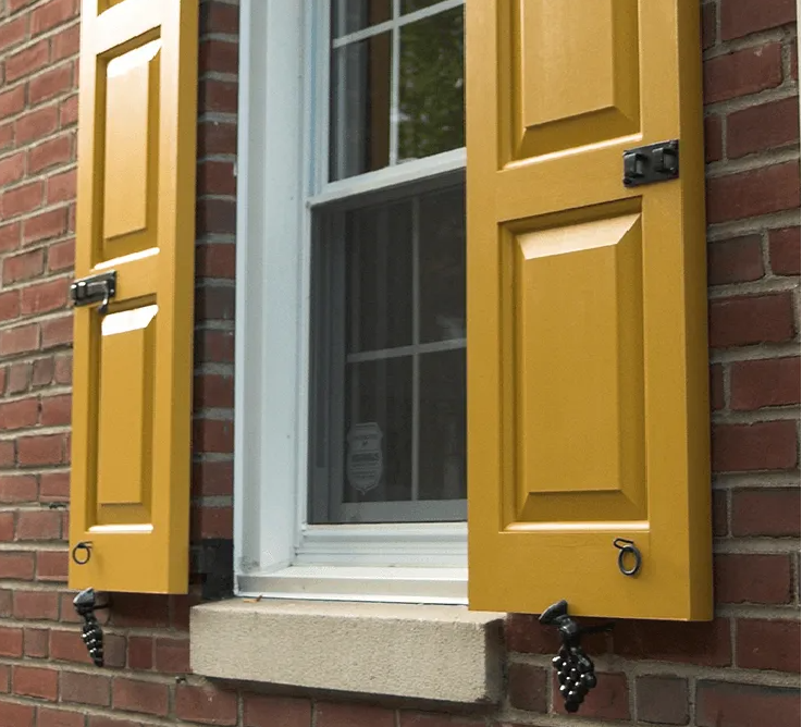Mustard yellow shutters with decorative black hardware mounted on a brick wall beside a white-framed window.