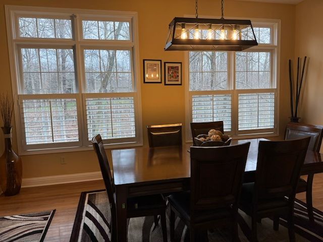 A dining room featuring a dark wood table, chairs, an industrial-style light fixture, and two large windows with shutters.