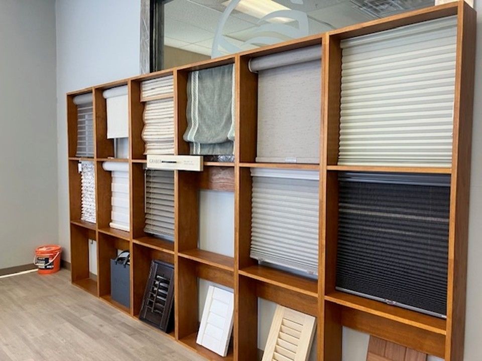 A wooden shelving unit displaying a variety of window shade samples, fabrics, and shutters in a showroom setting.