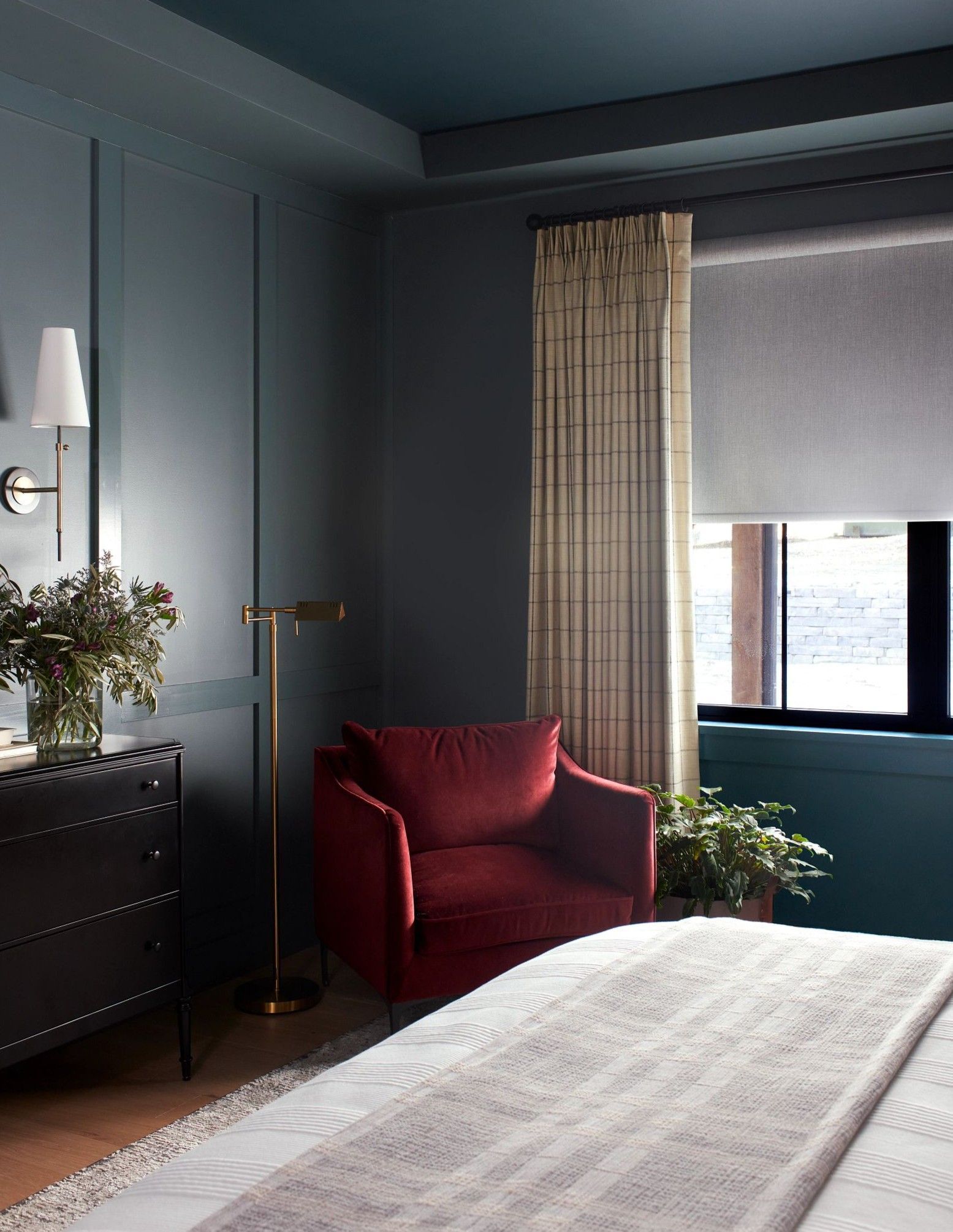 A dark teal bedroom with a deep red velvet armchair, a black chest of drawers, and a window with patterned curtains.