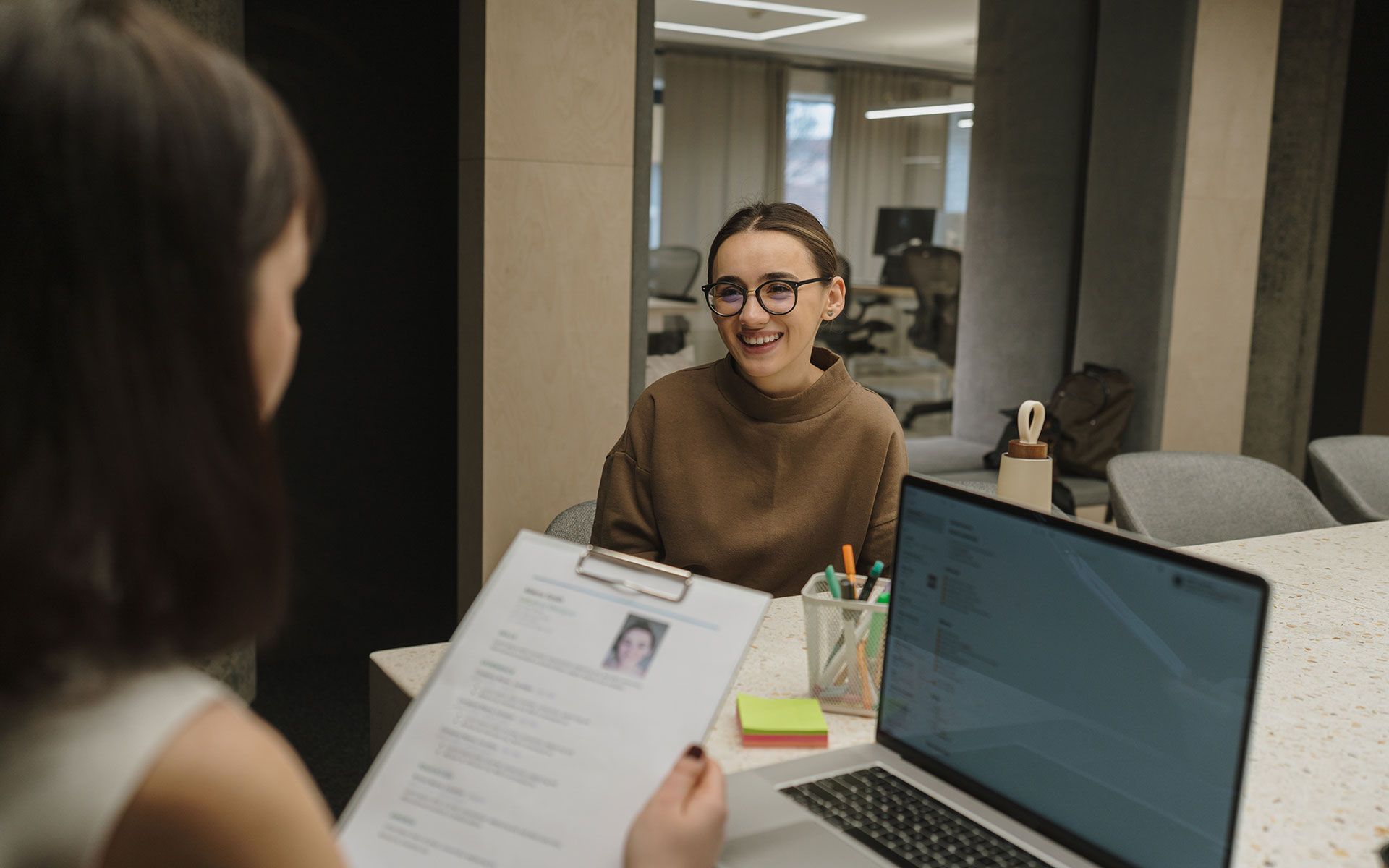 A woman is sitting at a table with a laptop and a clipboard.