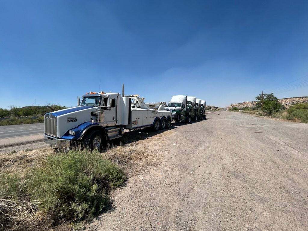 A tow truck pulling several semi-trucks on a dirt road, under a clear blue sky.