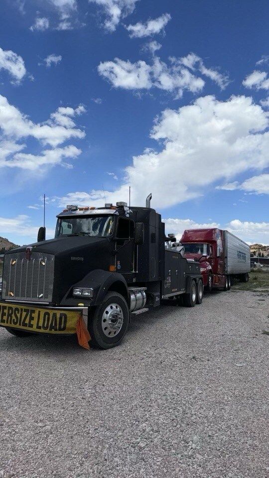 A tow truck is towing a semi truck in a gravel lot.