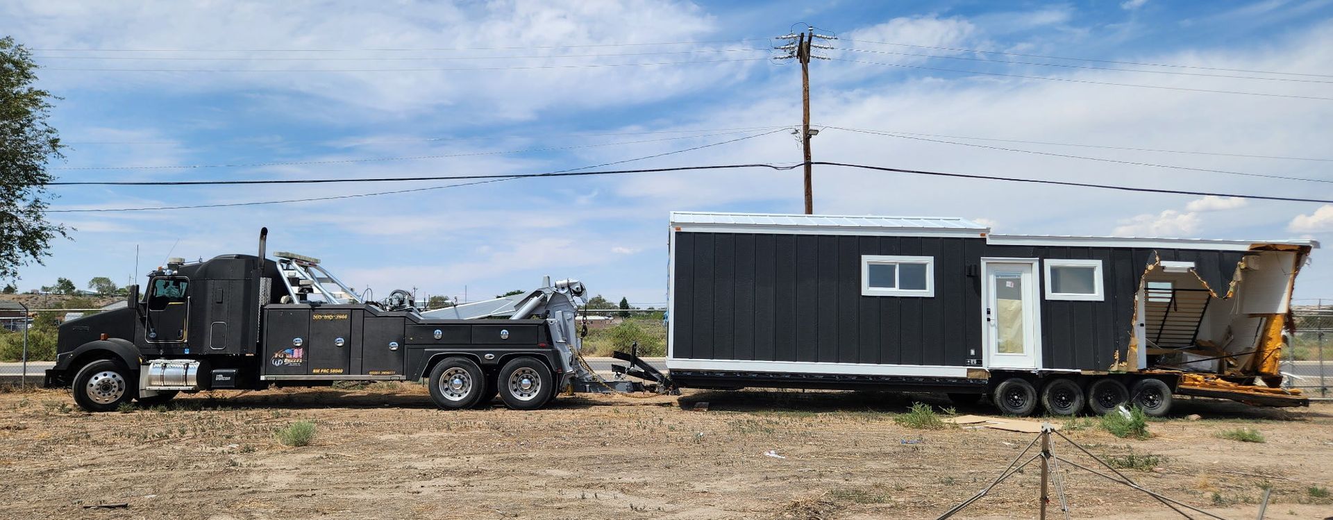 A black tow truck hauling a dark tiny house along a dirt road under a cloudy sky.