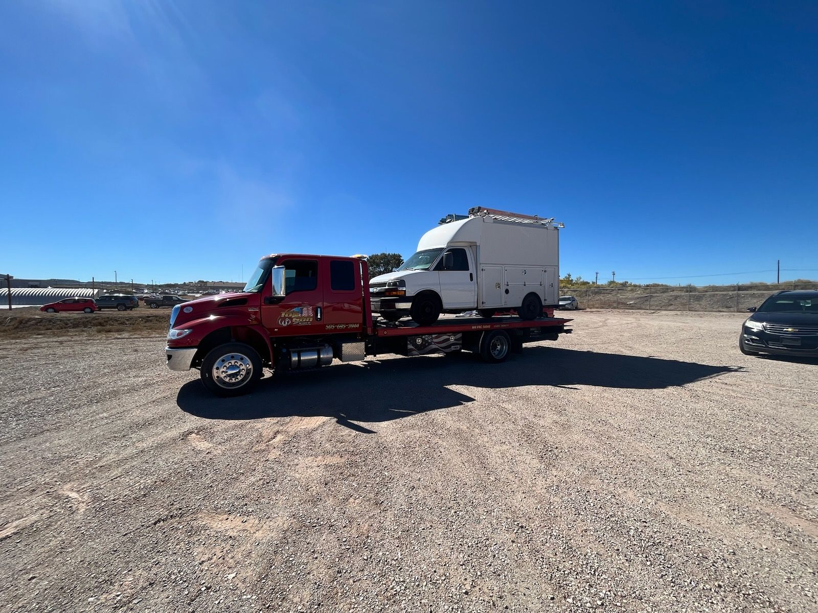A red tow truck is towing a white van in a gravel lot.