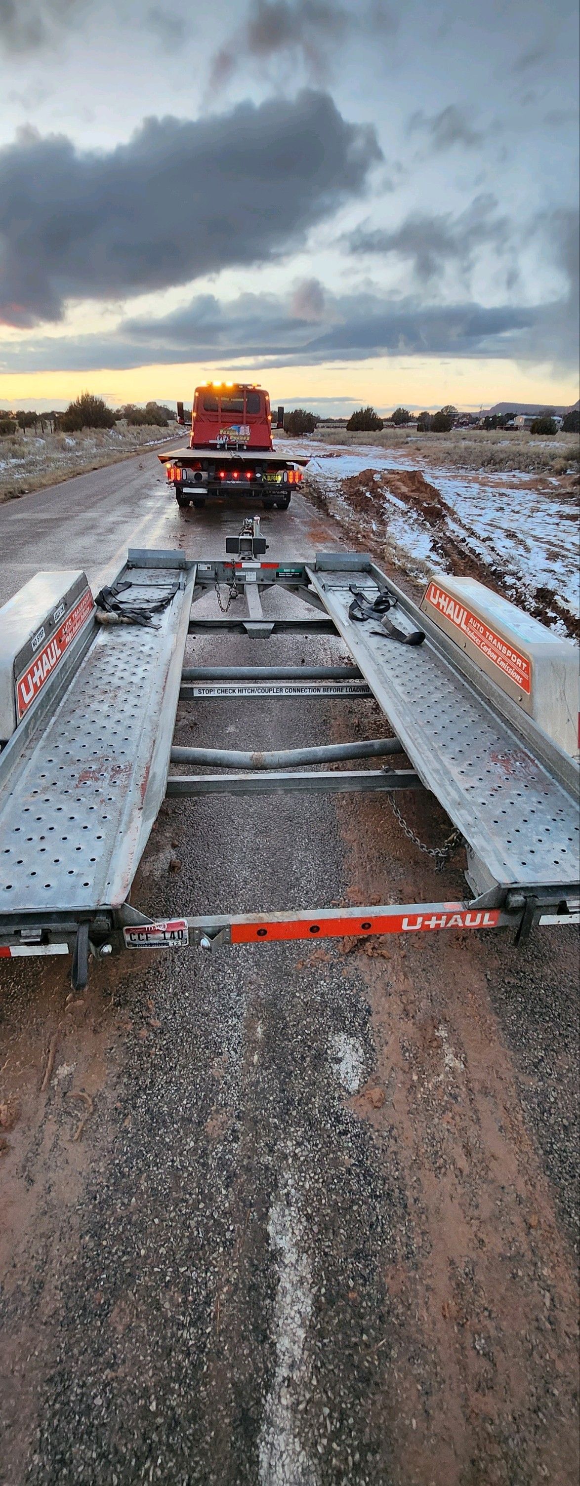 A tow truck is driving down a dirt road next to a trailer.