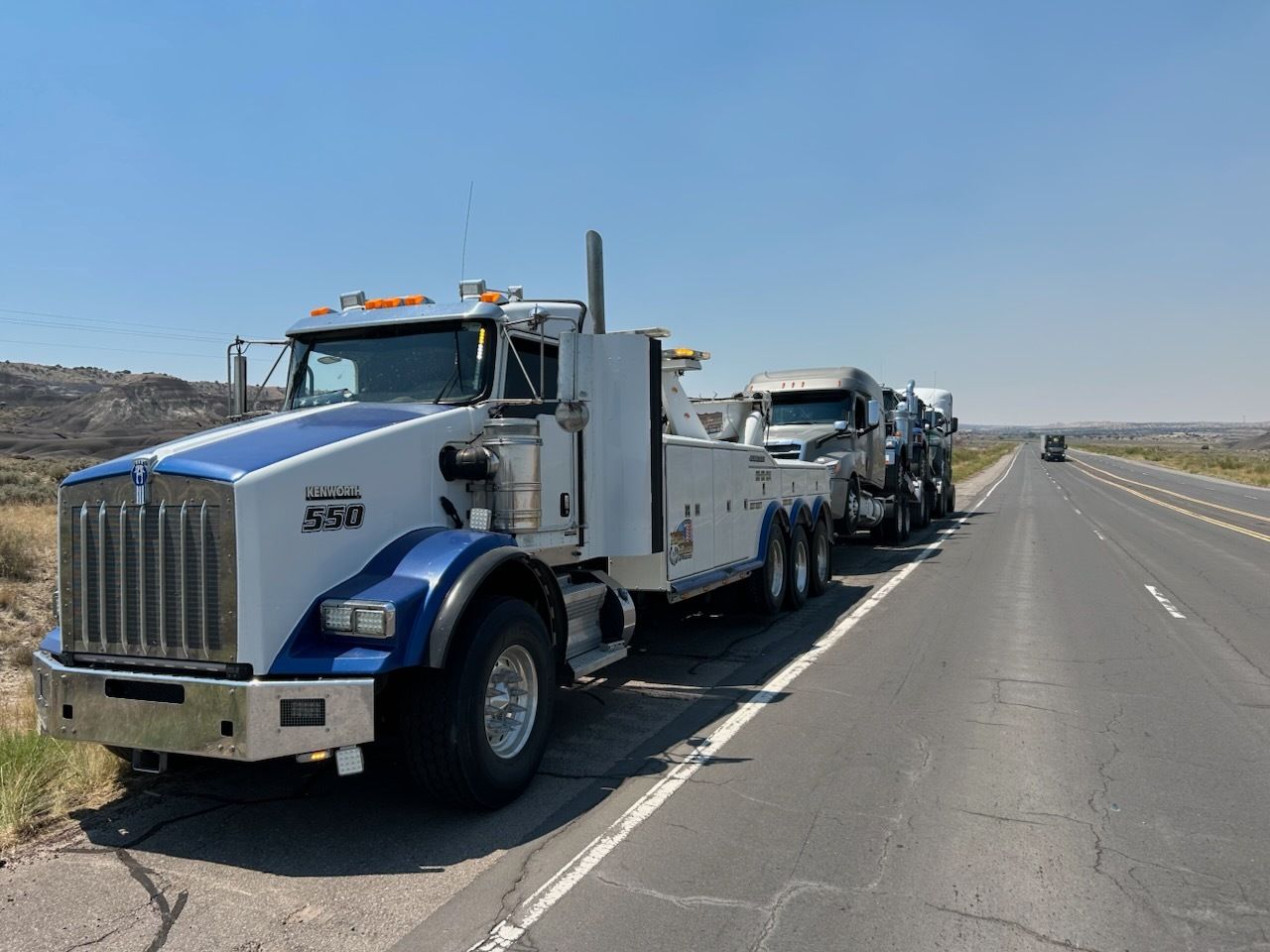 A large tow truck with a blue and white cab hauls two vehicles on a sunny highway.