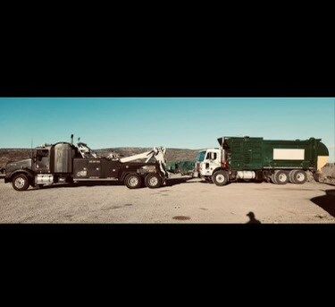 A tow truck is towing a garbage truck in a parking lot.