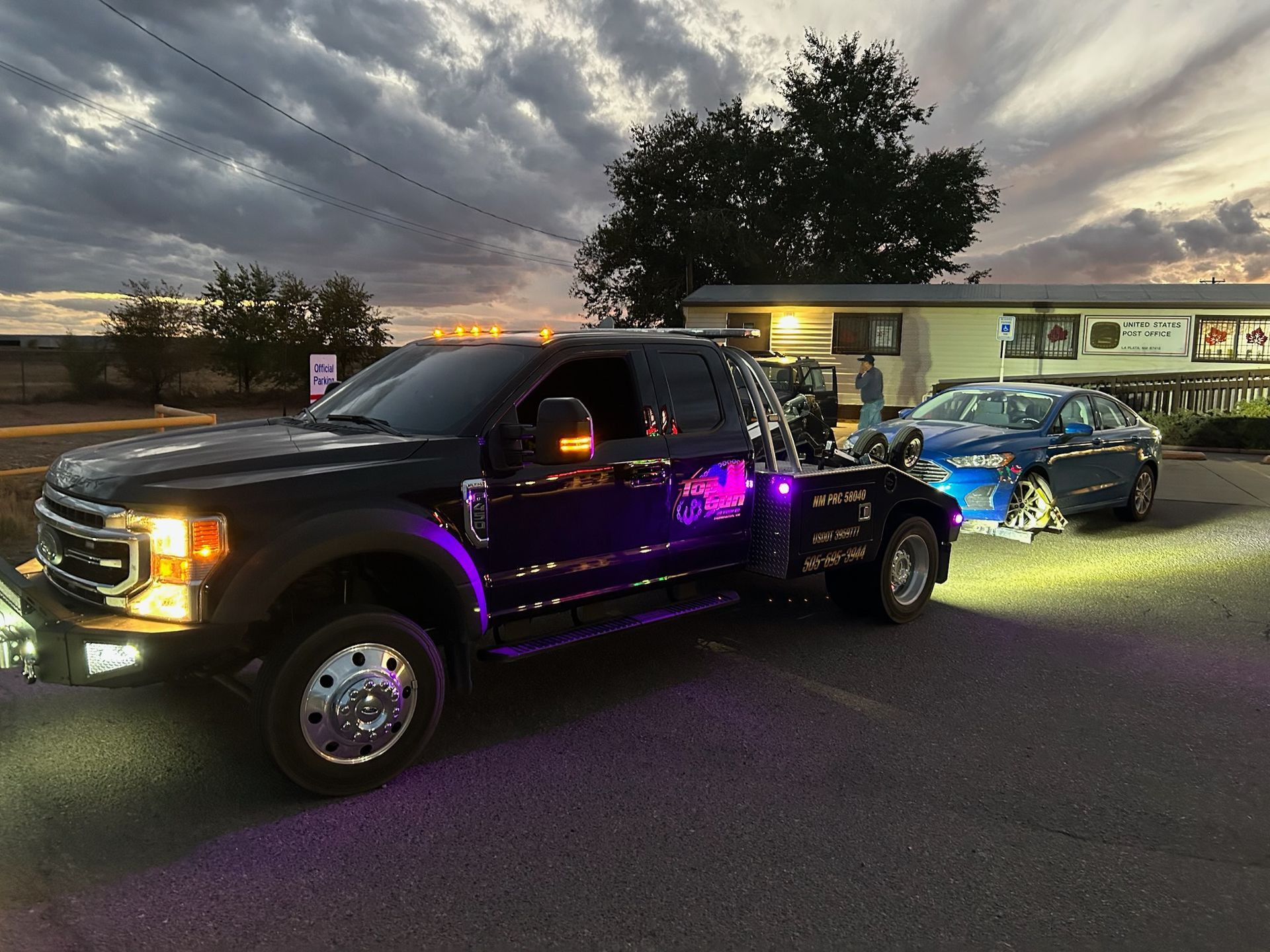 A black tow truck is towing a blue car in a parking lot.