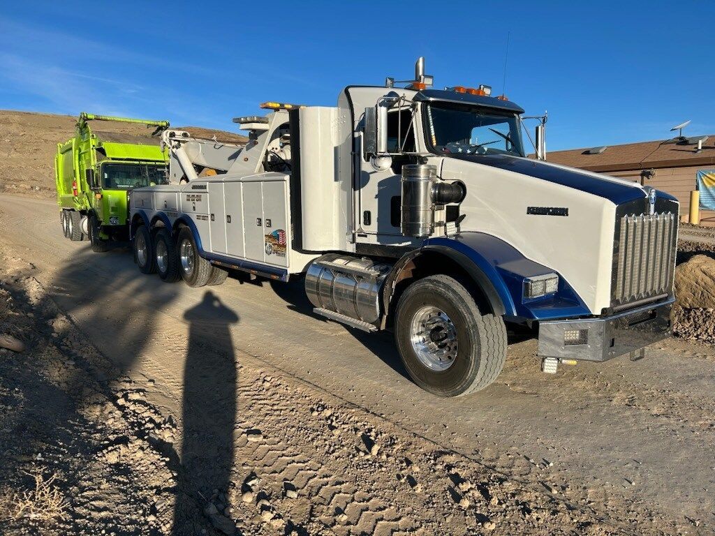A white tow truck is parked on the side of a dirt road next to a green dump truck.