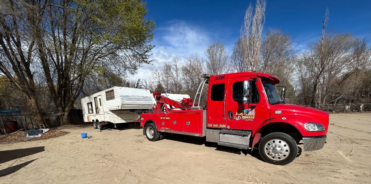 A red tow truck is towing a trailer in a dirt lot.