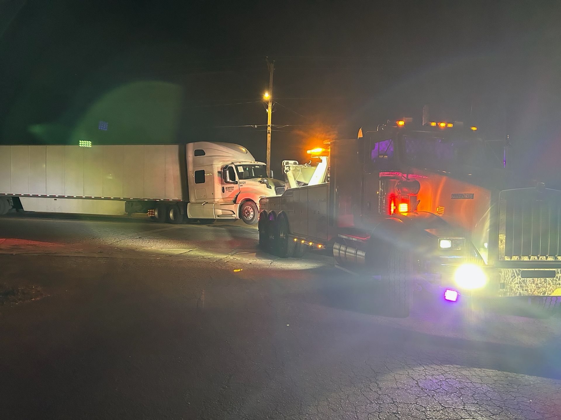 A semi truck and a tow truck are parked on the side of the road at night.