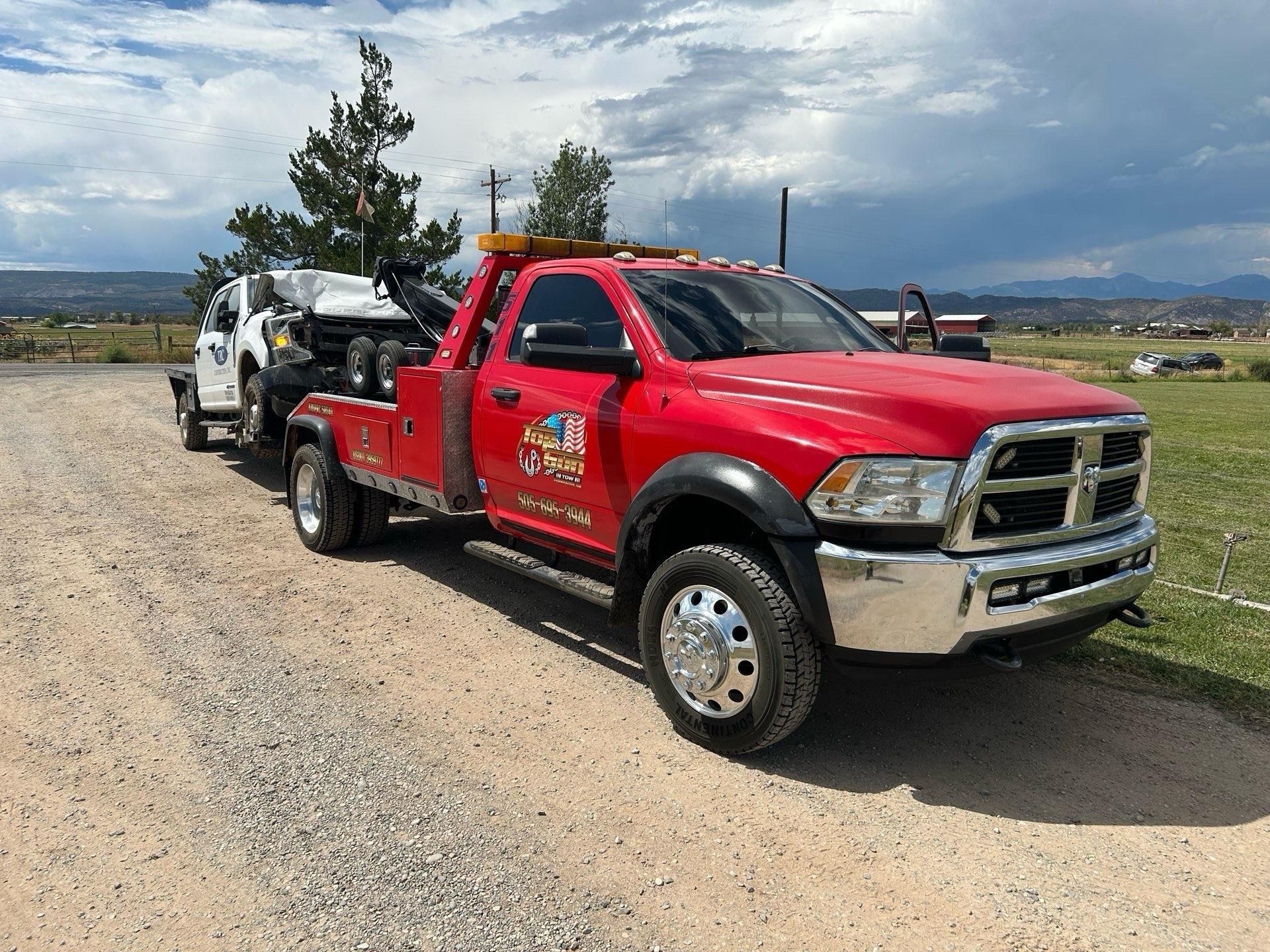 Red tow truck towing a white vehicle on a gravel road with mountains in the background under a cloudy sky.