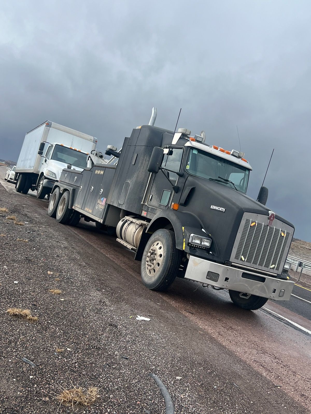 A black tow truck is driving down a gravel road next to a white truck.