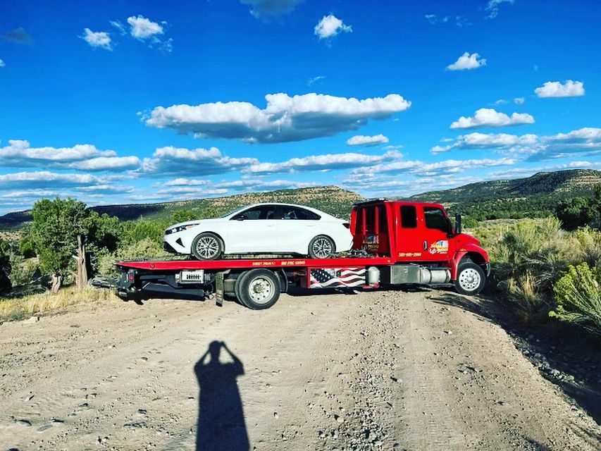 A tow truck is towing a white car on a dirt road.