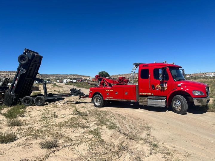 A red tow truck is towing a dump truck on a dirt road.